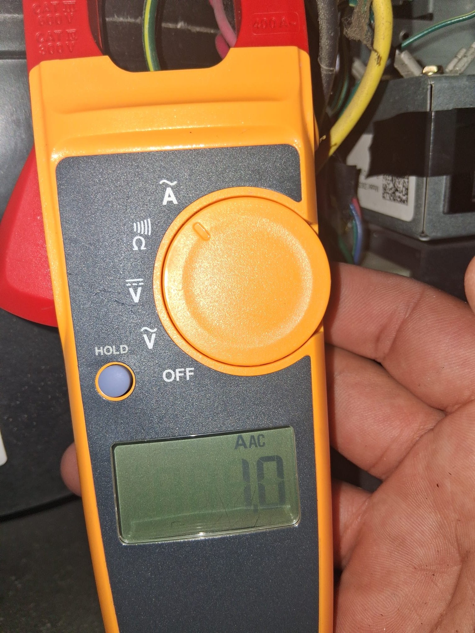 The image shows an HVAC technician using a clamp-style digital multimeter to measure AC amperage inside an HVAC unit’s electrical compartment.